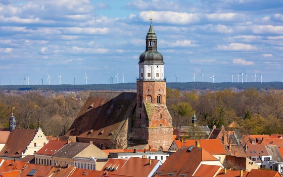 Kirchturm &uuml;ber roten D&auml;chern einer Stadt, dahinter Wald und Windr&auml;der unter blauem, wolkigem Himmel., Foto: Andreas Franke, Lizenz: Andreas Franke